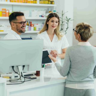 AdobeStock_475615519 Two pharmacist giving prescription medications to senior female customer in a pharmacy while using digital tablet